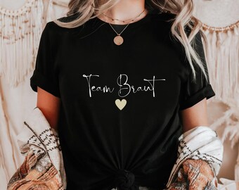 Może przedstawiać: Czarny T-shirt z białym tekstem, który brzmi "Team Braut" z małym żółtym sercem pod tekstem.