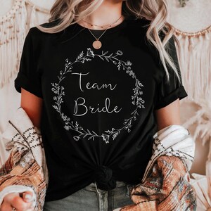 Op de afbeelding: Zwart T-shirt met een wit bloemkransontwerp en de tekst "Team Bride" in het midden.