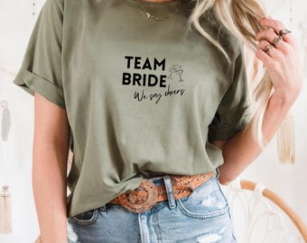 Może przedstawiać: Oliwkowo-zielona koszulka z czarnym napisem "TEAM BRIDE We say cheers". Projekt zawiera dwa kieliszki do szampana. Koszulka jest noszona z niebieskimi dżinsami i brązowym paskiem.
