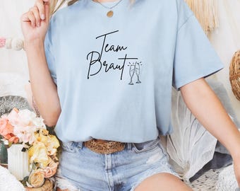 May include: A light blue t-shirt with the text "Team Braut" in black script and two champagne glasses with a sparkle above them.