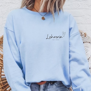 Könnte beinhalten: Hellblauer Pullover mit dem Text "Lehrerin mit Herz" und einem Herzsymbol.