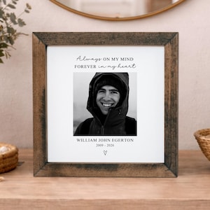 May include: A framed black and white photograph with the text "Always on my mind forever in my heart." The photo features a smiling person wearing a hat and jacket. The frame is made of dark brown wood.
