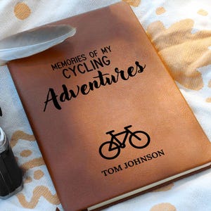 May include: A brown leather journal with the text "MEMORIES OF MY CYCLING ADVENTURES" and a bicycle graphic. The name "TOM JOHNSON" is printed at the bottom. A feather rests on the journal.