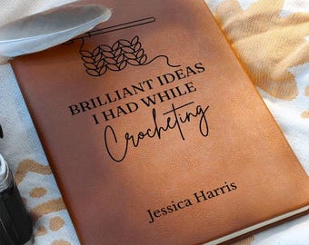 May include: A brown leather journal with the text "BRILLIANT IDEAS I HAD WHILE Crocheting" and the name "Jessica Harris". The cover features a crochet hook and yarn design. A feather rests on the journal.
