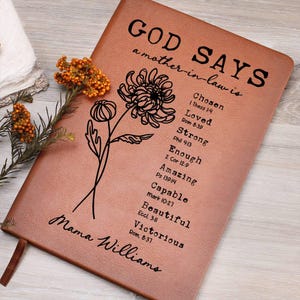 May include: A brown leather journal with the words "GOD SAYS a mother-in-law is" and a list of positive attributes. A floral illustration and the name "Mama Williams" are also featured. A sprig of orange flowers rests on the side.