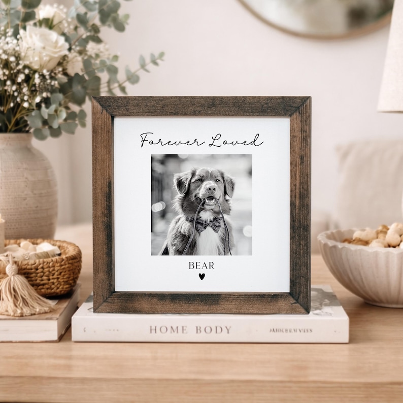 Puede incluir: Un marco de fotos cuadrado de madera con una imagen en blanco y negro de un perro. La foto tiene las palabras "Forever Loved" encima de la imagen y el nombre "Bear" debajo, con un s&iacute;mbolo de coraz&oacute;n. El marco est&aacute; sobre una pila de libros.