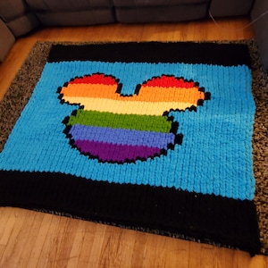 May include: A handmade, chunky knit blanket featuring a large, pixelated rainbow-colored Mickey Mouse head on a light blue background. The blanket has black borders and is made of soft, plush yarn.