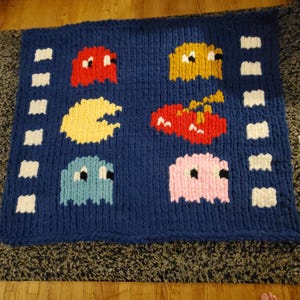 Peut inclure: Une couverture en tricot épais bleu marine, ornée de personnages pixélisés du jeu d'arcade classique Pac-Man. La couverture présente Pac-Man, des fantômes et le chemin du jeu en rouge, jaune, bleu et rose.