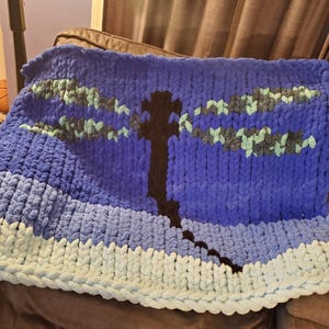 May include: A blue, green, and white knitted blanket with a black dragonfly silhouette in the center.