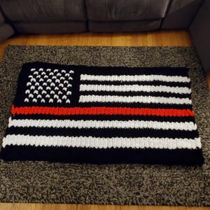 Puede incluir: Una manta rectangular hecha a mano con un diseño que se asemeja a la bandera estadounidense. La manta presenta un fondo negro con rayas blancas y una franja roja en el centro. La esquina superior izquierda tiene un campo de estrellas blancas sobre fondo negro.