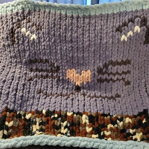 Puede incluir: Una manta de crochet de color morado con diseños de corazón y murciélago. La manta tiene un borde con estampado marrón y blanco.