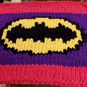May include: A handmade, chunky knit blanket featuring a Batman logo. The blanket has a purple center with a black and yellow Batman symbol. The top and bottom borders are bright pink, creating a bold contrast.