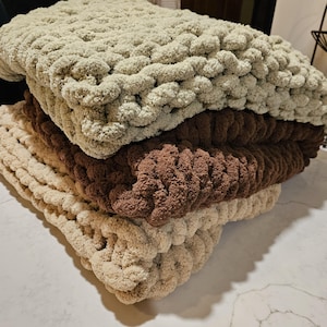 May include: A stack of four chunky knit blankets in varying shades of beige and brown. The blankets are folded neatly, showcasing the thick, textured yarn. The top blanket is a light beige, followed by a darker brown, and two other beige blankets.