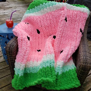 Può includere: Una coperta all'uncinetto rosa, verde e bianca con un motivo a cocomero. La coperta è drappeggiata su una sedia in vimini.