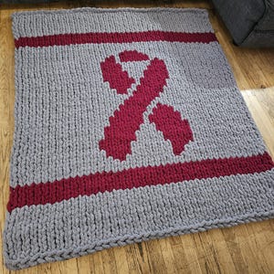 May include: A gray knitted throw blanket with a large red ribbon design. The blanket has a thick, chunky knit texture.