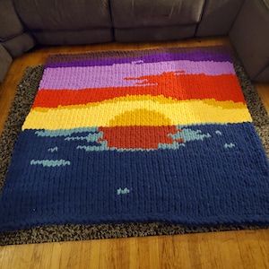 May include: A large, colorful knitted blanket with a sunset design. The blanket is made of thick yarn and has a variety of colors, including purple, red, yellow, and blue. The blanket is laid out on a wooden floor.