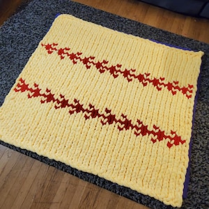 May include: A chunky knit blanket in a soft yellow hue, adorned with two diagonal rows of red heart-shaped patterns. The blanket is square and appears to be hand-knitted, offering a cozy and textured aesthetic. The blanket is resting on a dark gray surface.