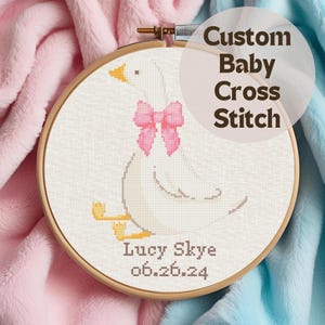 May include: A white goose cross stitch pattern with a pink bow, personalized with the name "Lucy Skye" and the date "06.26.24".