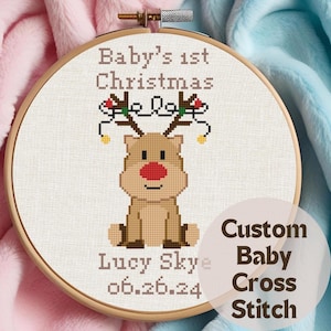 以下が含まれることがあります： 「Baby's 1st Christmas」の文字とトナカイのデザインが施されたクロスステッチのフレーム。トナカイは赤い鼻をしており、お祝いのライトで飾られています。「Lucy Skye」という文字と日付も含まれています。「Custom Baby Cross Stitch」の文字も表示されています。