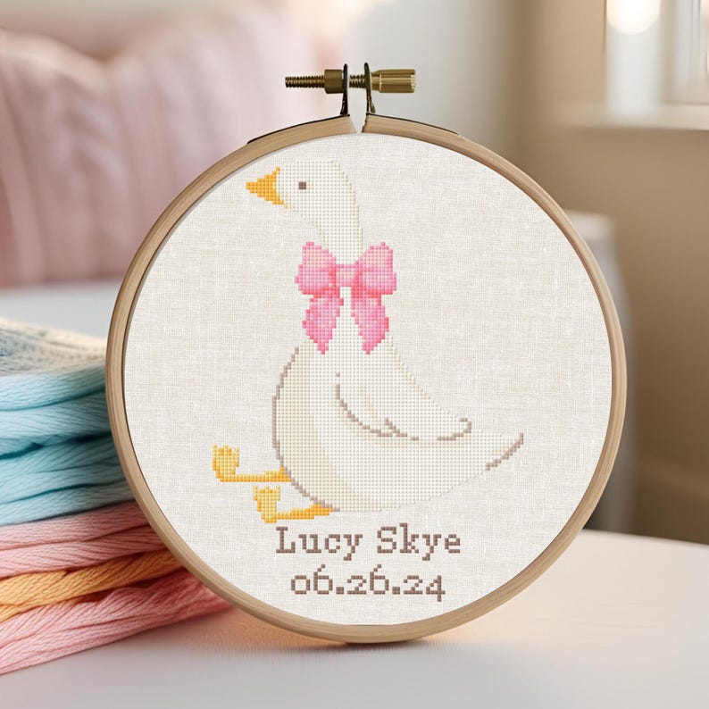 May include: A white embroidered goose with a pink bow, in a wooden embroidery hoop. The text "Lucy Skye 06.26.24" is embroidered below the goose.