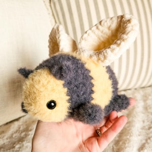 May include: A crocheted plush bee toy with yellow and gray fur, black eyes, and white wings.