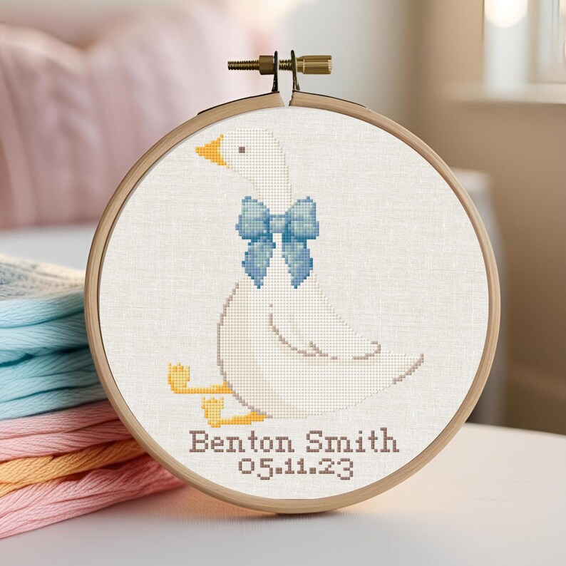 May include: A white embroidered goose with a blue bow, sitting on a white background. The embroidery is in a hoop and the text "Benton Smith 05.11.23" is below the goose.