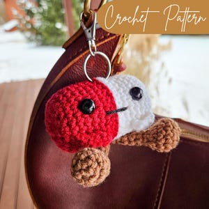 May include: A crocheted mushroom keychain with a red and white cap and brown stem. The keychain is attached to a silver key ring and is hanging from a brown leather bag.