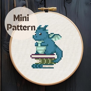 May include: A cross-stitch pattern of a blue dragon sitting on a stack of books, displayed in a wooden embroidery hoop. The text "Mini Pattern" is in a circular label above the dragon. The dragon has wings and is made of small, square stitches.