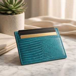 May include: A teal card holder with a textured lizard-skin pattern featuring two slots. One slot contains a solid black card, and the other slot contains a solid gold-colored card. The card holder is rectangular with stitched edges.