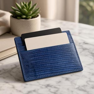 May include: A blue card holder with a textured lizard-skin pattern featuring two slots. One slot contains a solid black rectangular card, and the other slot contains a solid beige rectangular card. The card holder has visible stitching along the edges.