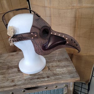 May include: A brown leather plague doctor mask with a long beak and eye holes. The mask is decorated with metal rivets and has a strap for wearing.