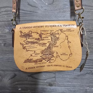 May include: A tan leather crossbody bag with a map design and the text "A DRAGON WITHOUT ITS RIDER IS A TRAGEDY" and "A RIDER WITHOUT THEIR DRAGON IS DEAD". The bag has a long, adjustable strap and metal hardware.