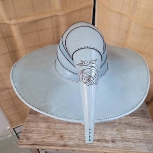 Puede incluir: Un sombrero de cuero azul claro con una corona alta y puntiaguda y un ala ancha. El sombrero tiene un diseño de rosa tallado en la parte delantera.