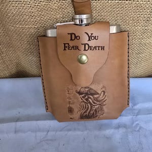 以下が含まれることがあります： ステンレス製のボディと革ストラップが付いた、茶色の革製フラスコ。フラスコの革ケースには「Do You Fear Death」の文字と生き物のイラストが描かれています。フラスコは液体を入れるように設計されています。
