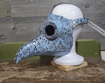May include: A light blue plague doctor mask with a floral pattern in brown. The mask has a long beak, large eye holes, and a strap for securing it. The mask is displayed on a white mannequin head.