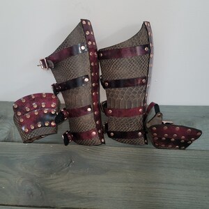 May include: A pair of leather arm and leg armor with a snakeskin pattern. The armor is primarily brown with burgundy accents and copper rivets. The design includes straps and buckles for fastening, suggesting a medieval or fantasy theme.