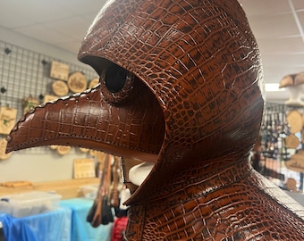 May include: A brown leather plague doctor mask with a long beak and a hood. The mask is made to look like crocodile skin.