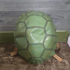 May include: A green turtle shell-shaped backpack with black stitching and brown leather straps. The backpack has a textured, reptile-like pattern and a rounded shape. The straps are attached to the shell with rivets.