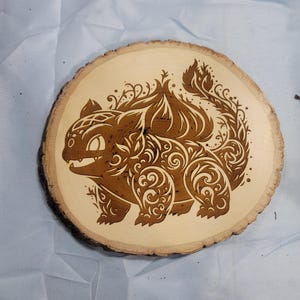 Könnte beinhalten: Eine runde Holzscheibe mit einem braunen, eingebrannten Design einer mythischen Kreatur mit filigranen Details. Die Kreatur hat einen großen Kopf mit einem breiten Maul und einen langen Schwanz.