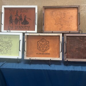 May include: Five wooden trays with leather inlays. The trays feature different designs, including a group of figures, a map, a dice, and a graphic. The trays are displayed on a blue surface.