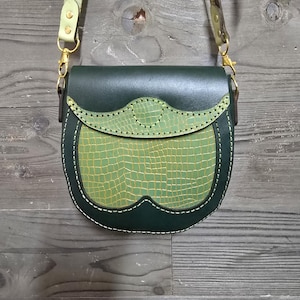Può includere: Una borsa a tracolla in pelle verde scuro con un pannello frontale verde chiaro con texture di alligatore. La borsa ha una tracolla verde chiaro abbinata e hardware color oro. Le cuciture bianche delineano il pannello frontale e i bordi della borsa.