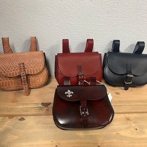 May include: Four leather bags with buckles and straps. The bags are in different colors: brown, red, black, and dark brown. The dark brown bag has a decorative metal fleur-de-lis.