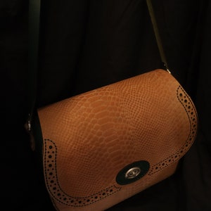 Puede incluir: Un bolso de cuero marrón cruzado con ribete verde y negro. El bolso tiene un patrón texturizado, similar a la piel de serpiente, y un cierre de metal decorativo.