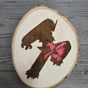 Puede incluir: Una rodaja redonda de madera con una silueta marrón de una figura con una silueta rosa de un pájaro dentro de la figura.