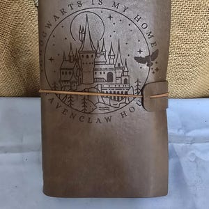 Puede incluir: Diario de cuero marrón con un escudo de Hogwarts que presenta un castillo, una luna y estrellas. El texto dice "Hogwarts es mi hogar Ravenclaw House".