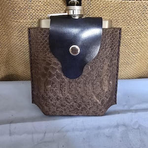 May include: A brown leather flask with a black leather flap and a snap closure. The flask has a snakeskin pattern.