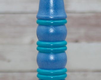 Puede incluir: Un taco de madera azul y turquesa con la parte superior e inferior redondeadas. El taco está pintado en un degradado de tonos azules con bandas turquesas.