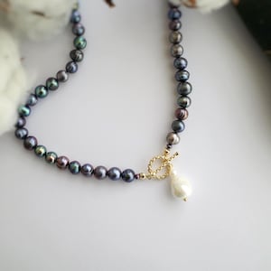 May include: A necklace featuring a strand of dark, iridescent, round pearls with a gold-toned toggle clasp. A single, larger, white, teardrop-shaped pearl hangs from the center of the clasp, creating a focal point for the jewelry piece.