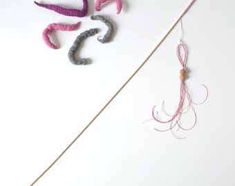 May include: A wooden stick with a pink and white string attached to the end. The string has two wooden beads and is tied in a knot. There are five felt shapes in the background, two pink, two gray, and one purple.