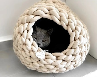 Pode incluir: Uma cama redonda para animais de estimação na cor creme, feita de tecido grosso e trançado. A cama possui uma grande abertura circular com um interior escuro onde um gato cinza descansa. Uma pequena etiqueta branca em forma de coração com o texto PETLOVEDE está fixada na parte frontal.